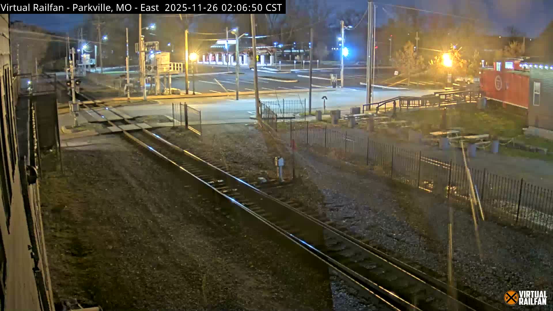 Train tracks cross a road at a railway crossing, flanked by a building on the left and a red caboose-like structure with outdoor seating on the right, all brightly illuminated by streetlights on a dark night.
