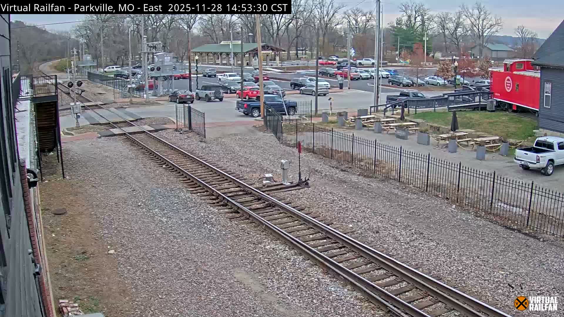 An overcast day reveals a scene with railroad tracks in the foreground, a busy road or parking lot with numerous cars, a red caboose repurposed next to a building, and an outdoor dining area with picnic tables.