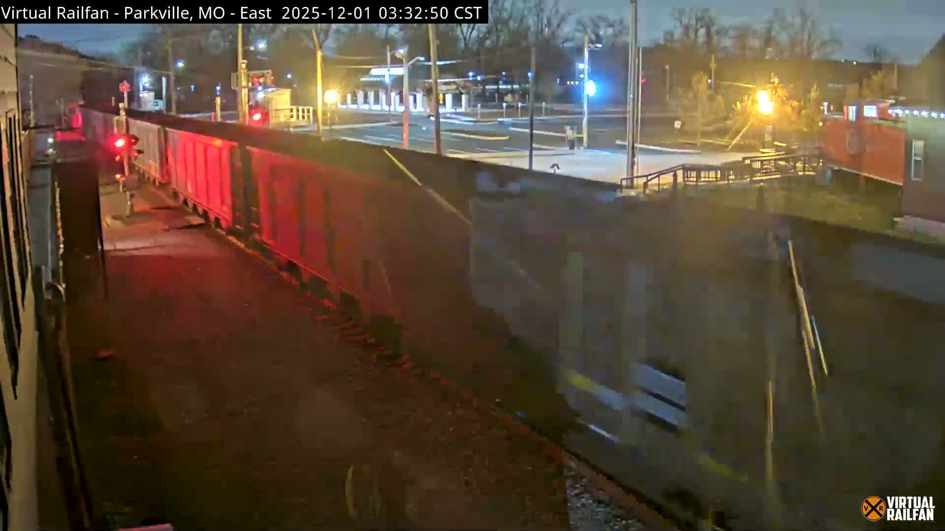 At night, a freight train moves past a railroad crossing with prominent red signals and streetlights in an illuminated urban area under an overcast sky.