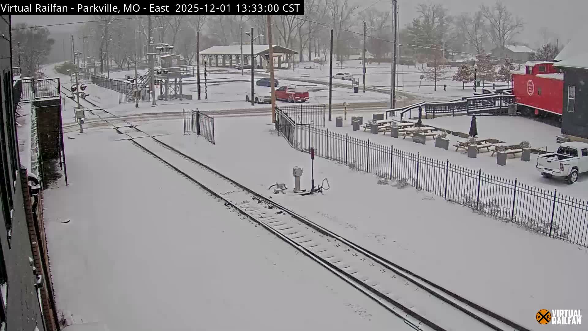 Snow-covered railroad tracks extend through a wintry landscape featuring a park or town square with a red caboose, outdoor seating, and parked vehicles, all blanketed under fresh snow on an overcast day.