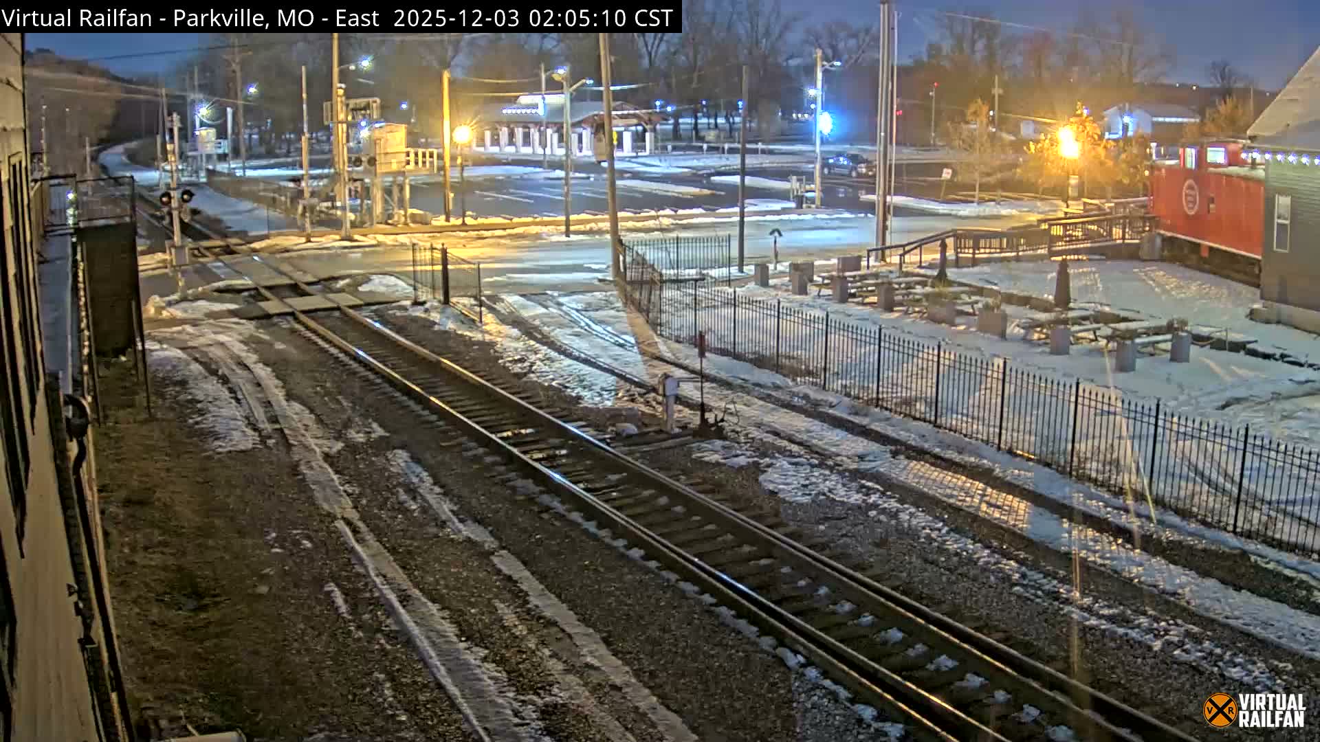 The image depicts a snowy railroad crossing with multiple tracks, illuminated by streetlights on a clear winter night, featuring a red train car and various buildings in the background.