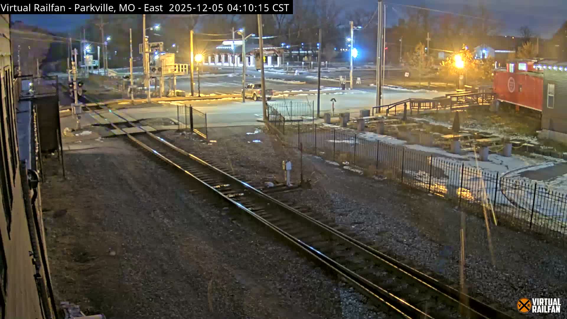 A nighttime view of a railroad crossing with multiple tracks, a pickup truck on the road, and patches of snow on the ground, all illuminated by streetlights, with a red caboose and outdoor seating visible in the background under clear skies.