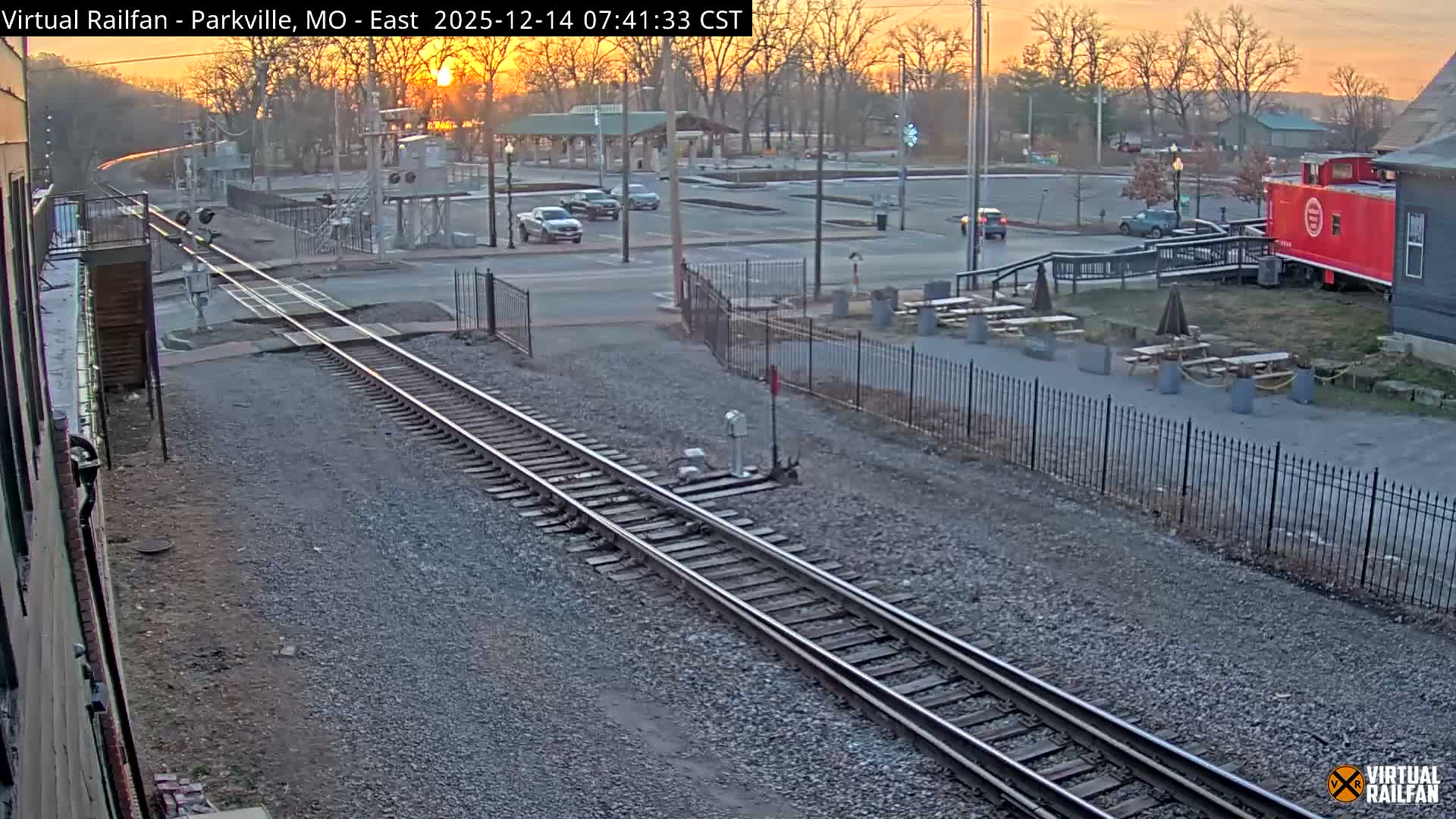 A nighttime view of a railroad crossing with multiple tracks, a pickup truck on the road, and patches of snow on the ground, all illuminated by streetlights, with a red caboose and outdoor seating visible in the background under clear skies.