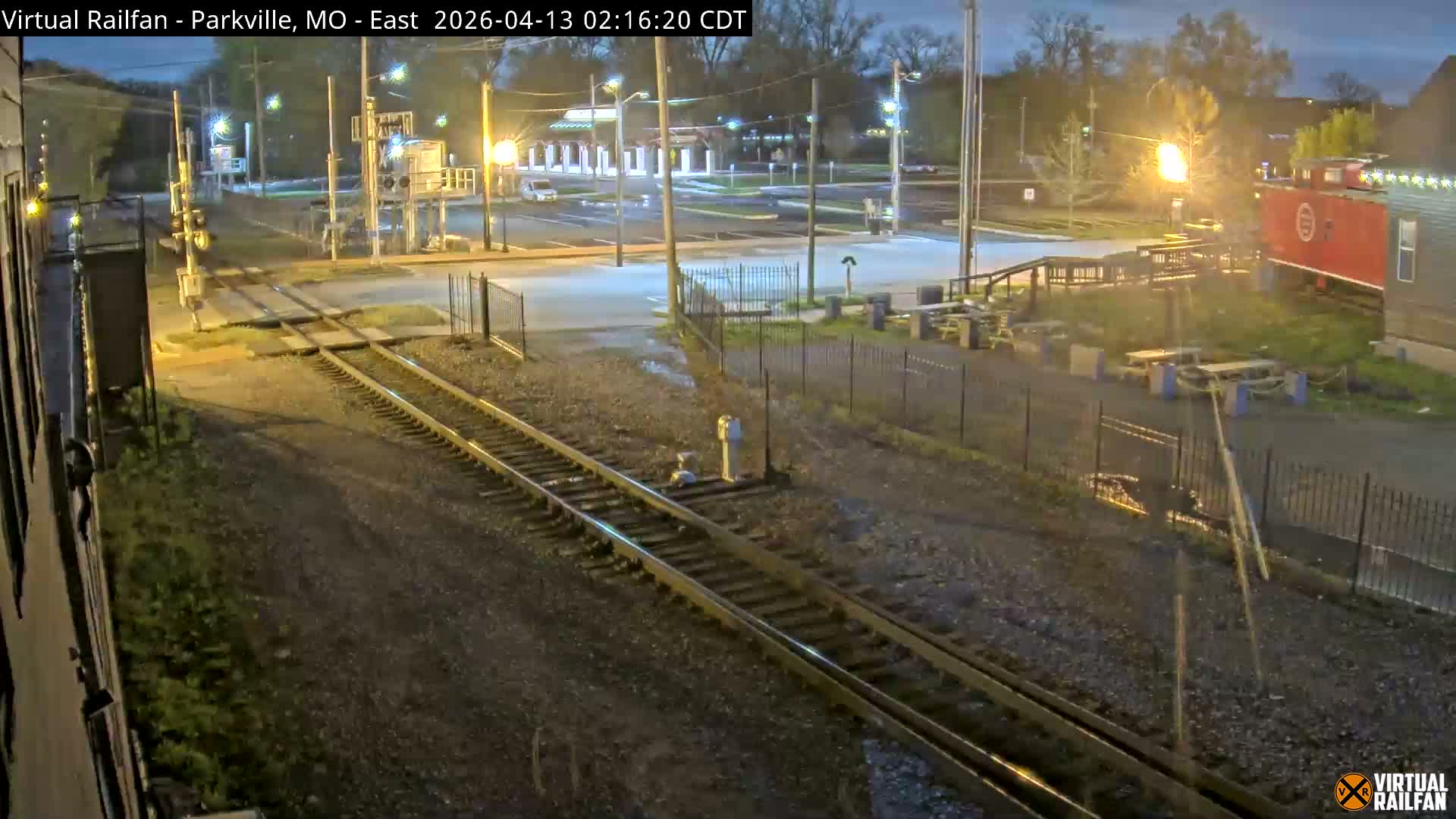 A nighttime view of a railroad crossing with multiple tracks, a pickup truck on the road, and patches of snow on the ground, all illuminated by streetlights, with a red caboose and outdoor seating visible in the background under clear skies.
