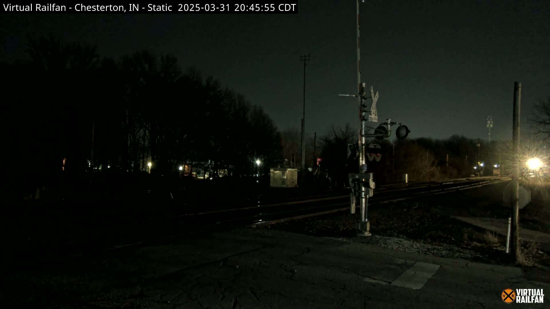 A nighttime view of a railroad crossing with train tracks extending into a dark, tree-lined area under a clear sky.