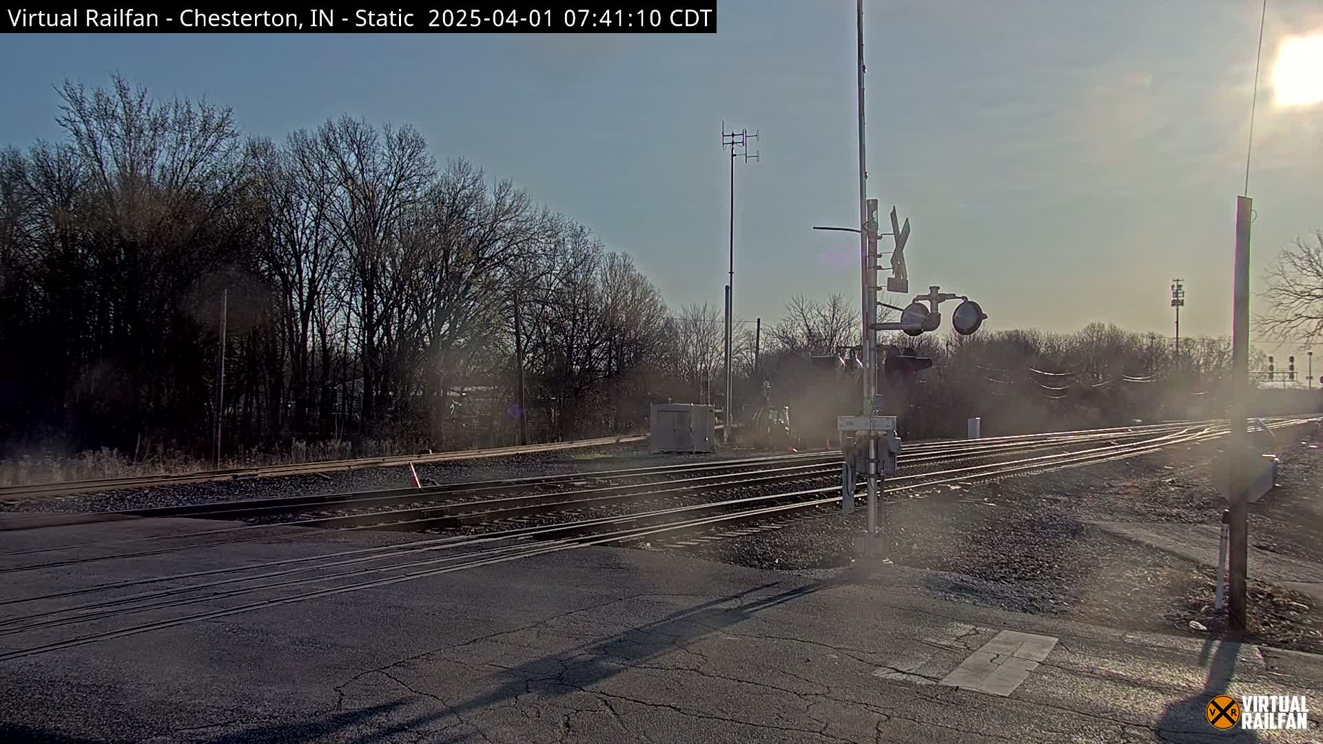A sunny day reveals a railroad crossing with multiple tracks, bare trees in the background, and train signal equipment.