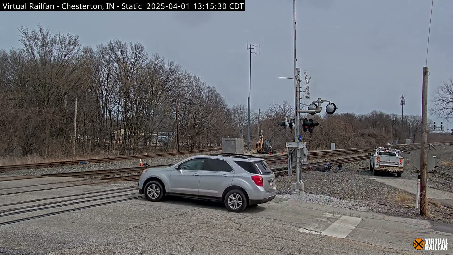 A silver SUV is stopped at a railroad crossing under an overcast sky, with bare trees and a work truck visible in the background.