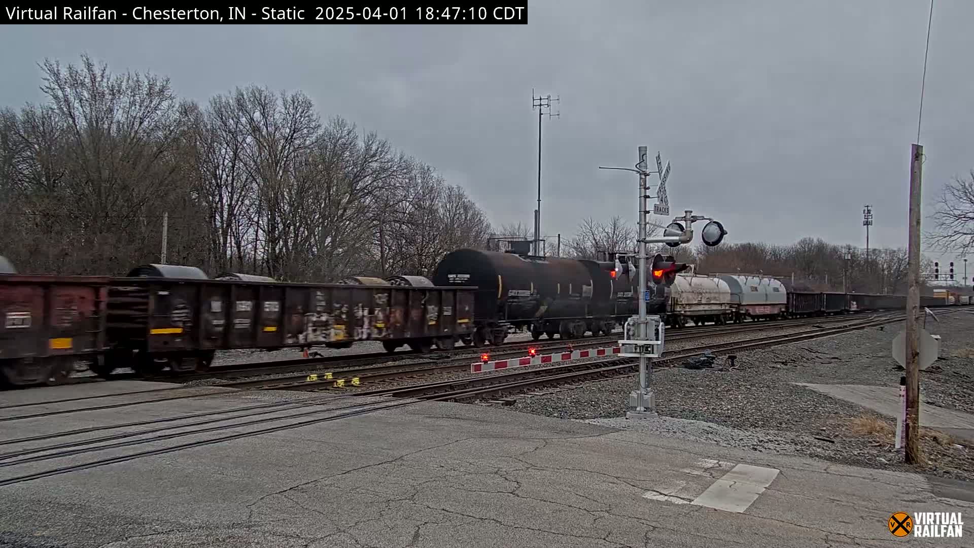 A long freight train passes a railroad crossing under an overcast sky.