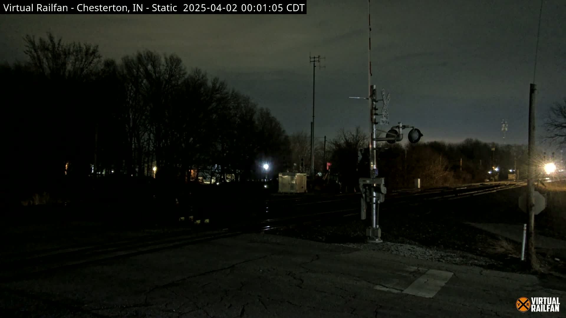 A nighttime view of a railroad crossing with a train signal, silhouetted trees, and dimly lit surroundings.