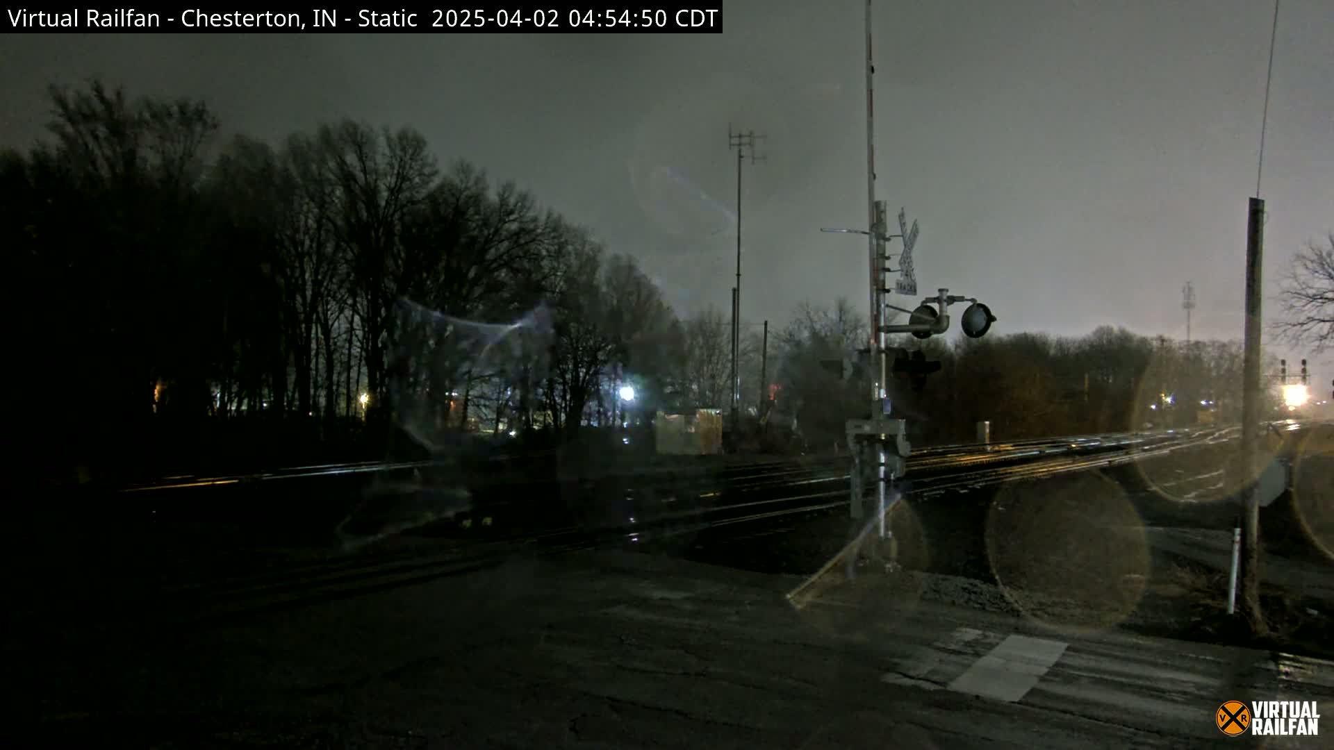 A nighttime view of a railroad crossing under a dark, overcast sky, with bare trees lining the background.