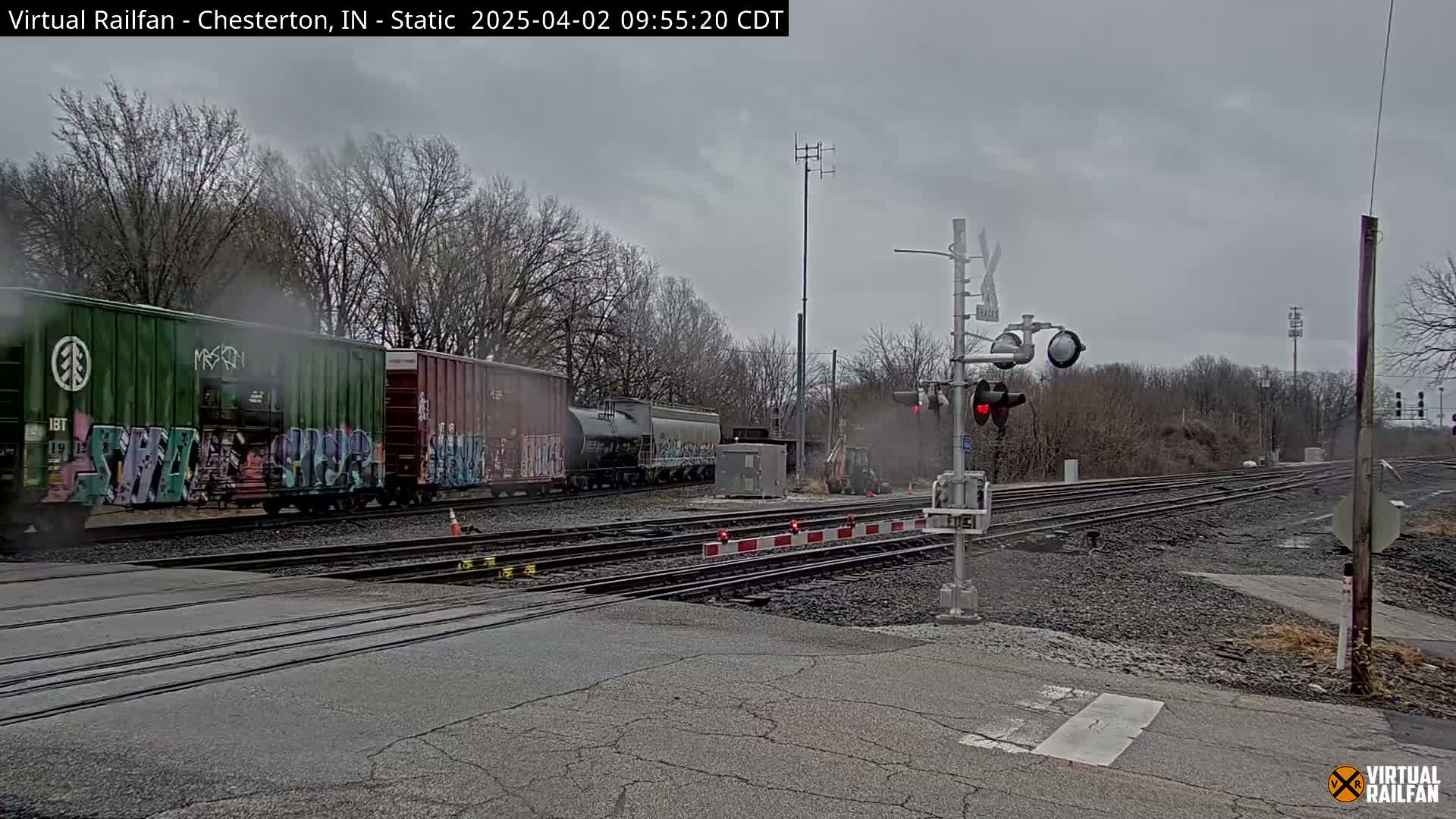 A freight train with graffiti-covered cars passes a railroad crossing under an overcast sky.