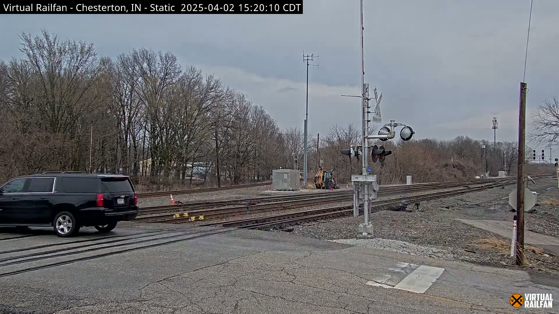 A black SUV drives across a railroad crossing under an overcast sky, past railroad signals and tracks, with bare trees lining the area.