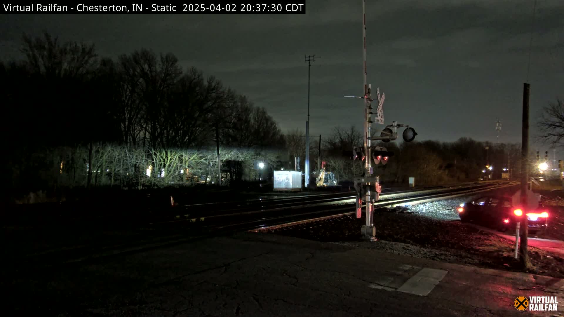 A nighttime view of a railroad crossing with a car stopped at the tracks under a dark, overcast sky.