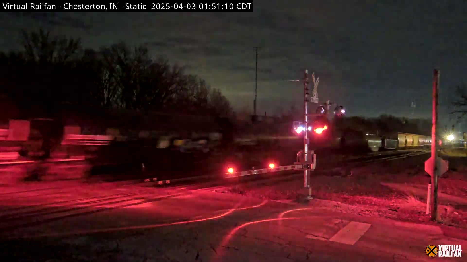 At night, a long freight train passes a road crossing with flashing red lights and lowered gates under a mostly clear sky.