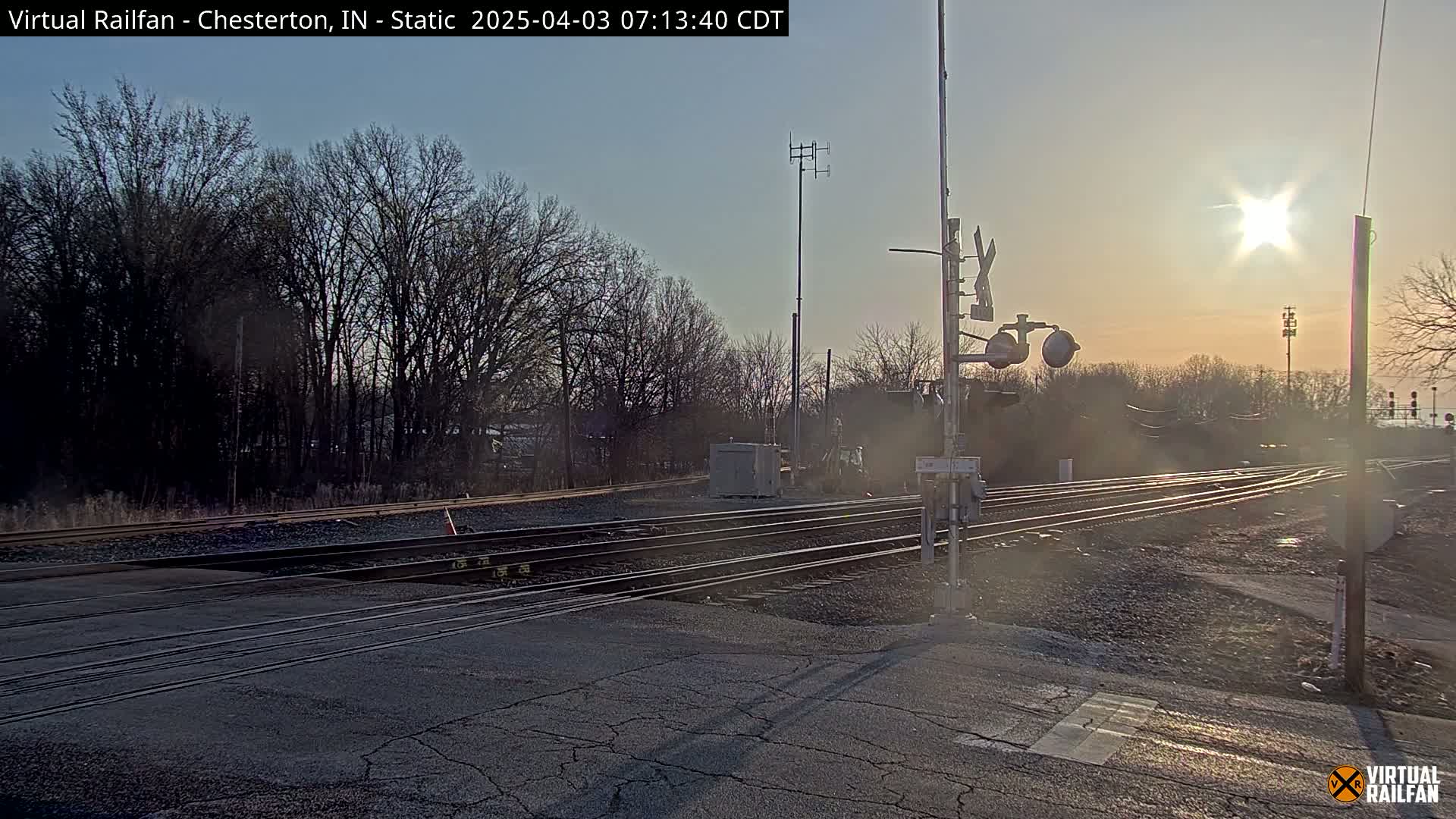 A railroad crossing with train tracks and signal lights is shown at sunrise under a mostly clear sky.