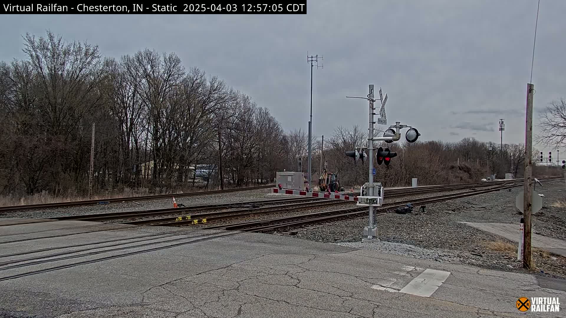 A railroad crossing with several tracks and a signal system is shown under an overcast sky.