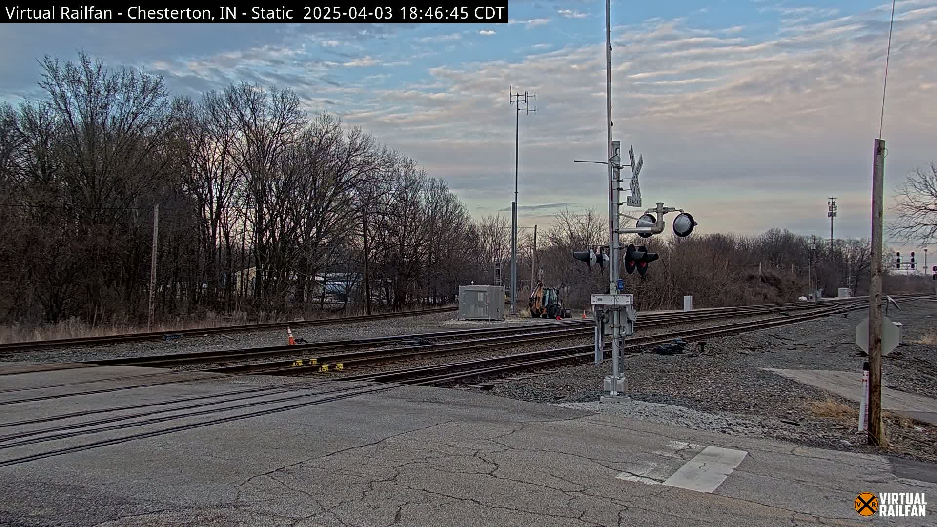A railroad crossing with a railway signal, bare winter trees, and a partly cloudy twilight sky.