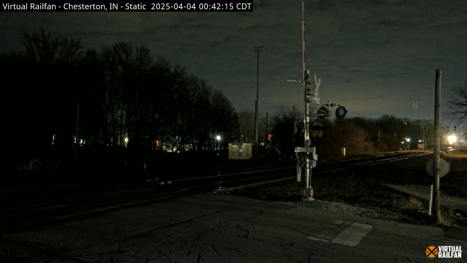 A nighttime view of a railroad crossing with train tracks extending into a dark, tree-lined area under a mostly clear, dark sky.