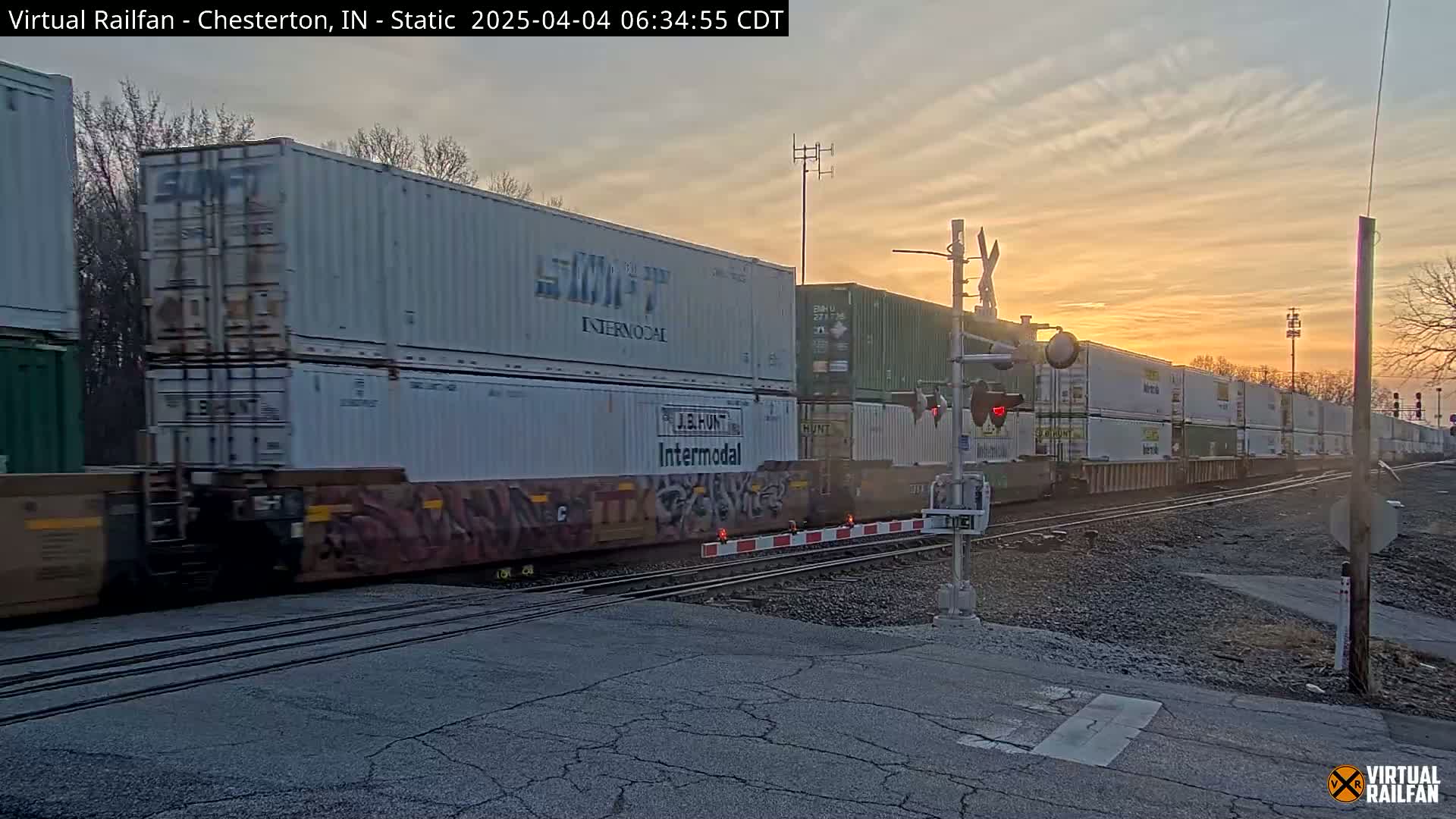 A long freight train carrying numerous white and green shipping containers passes a railroad crossing at sunset.
