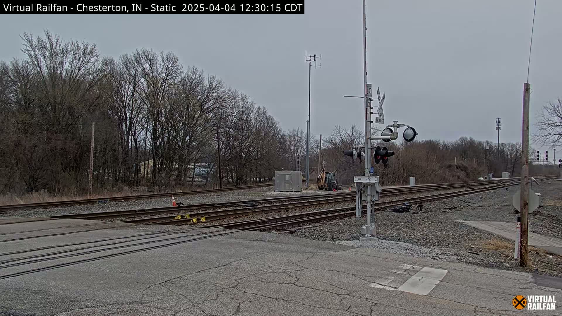A view of a railroad crossing with multiple tracks, under a cloudy sky, showing bare trees in the background and a small piece of construction equipment near the tracks.
