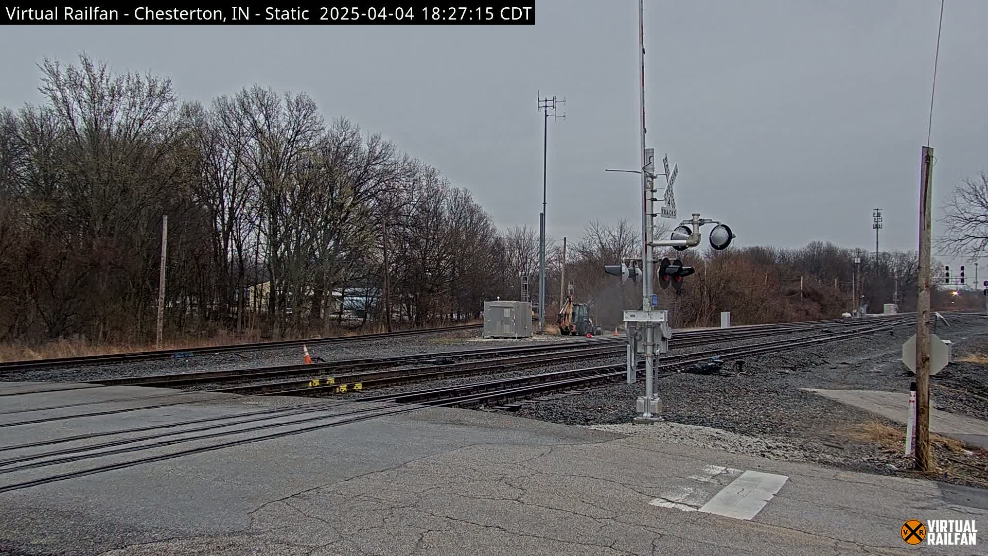 A railroad crossing with multiple tracks is shown under an overcast sky, with a small piece of construction equipment visible near the tracks and bare trees in the background.