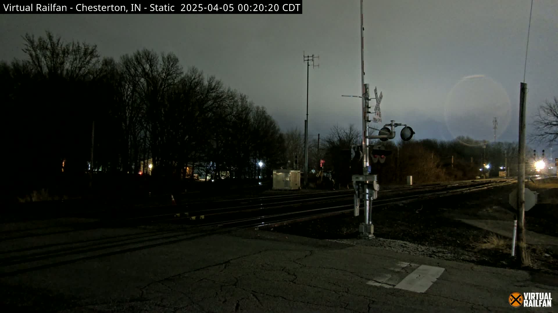 A nighttime view of a train crossing with a dark sky, bare trees, and railroad tracks extending into the distance.