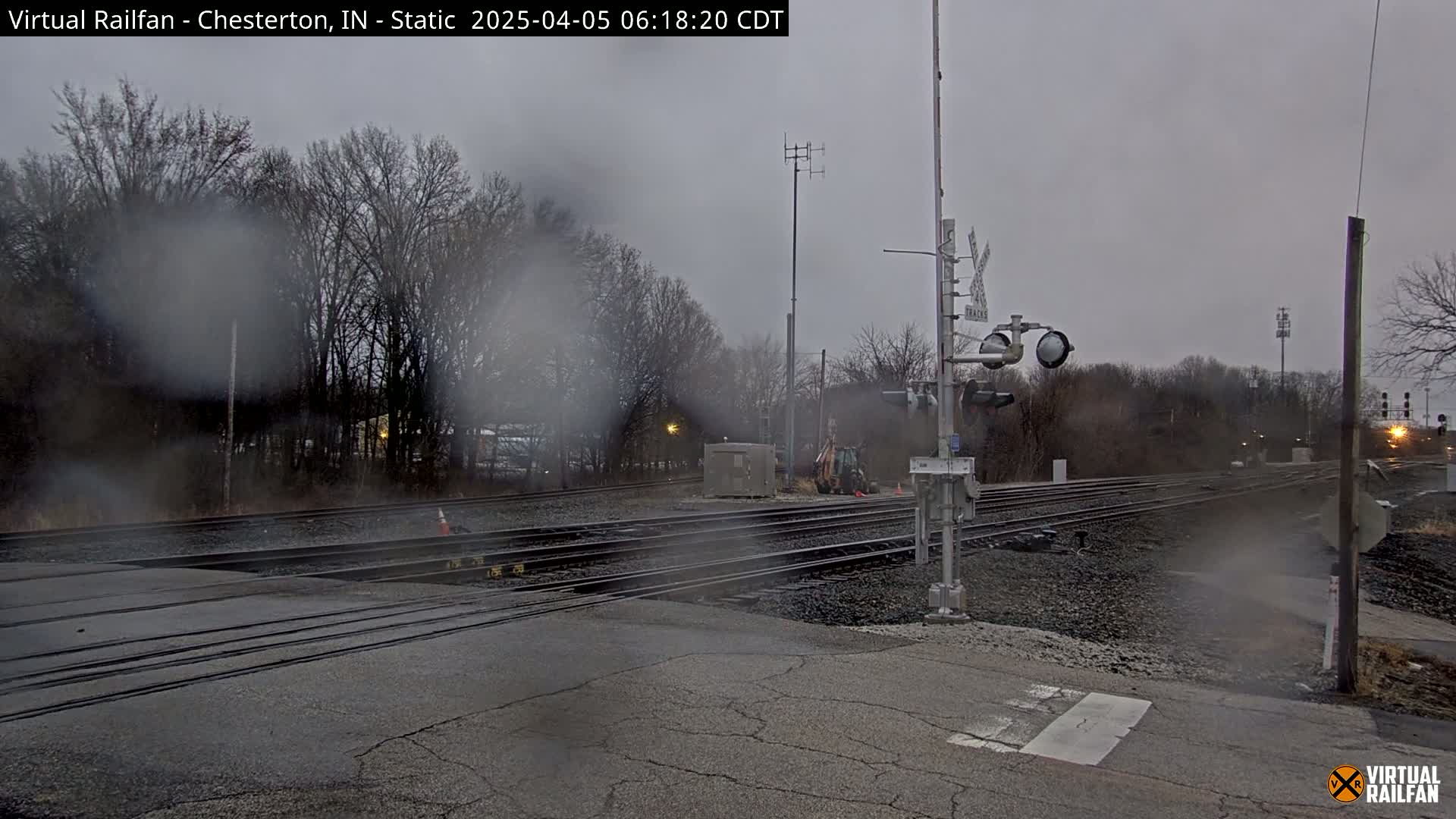 A rainy view of a railroad crossing with multiple tracks, surrounded by bare trees under an overcast sky.