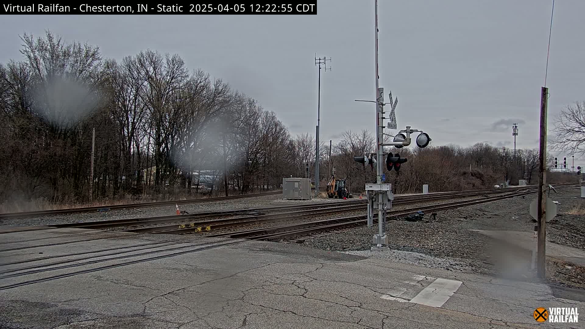 A rain-spattered view of a railroad crossing with multiple train tracks, crossing signals, and bare trees under an overcast sky.