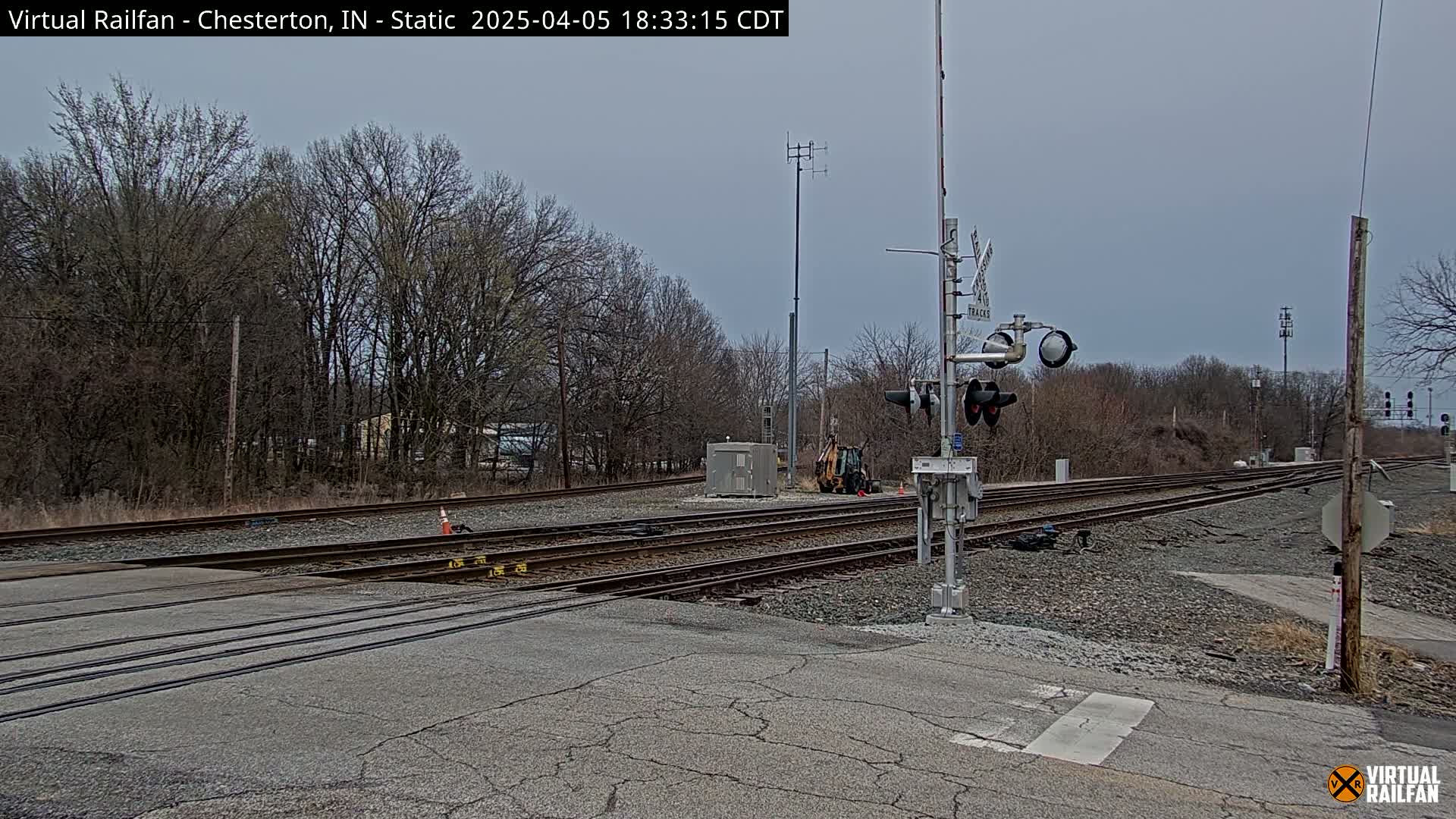 A railroad crossing with multiple tracks under a cloudy sky, bordered by bare trees and a small piece of construction equipment visible near the tracks.