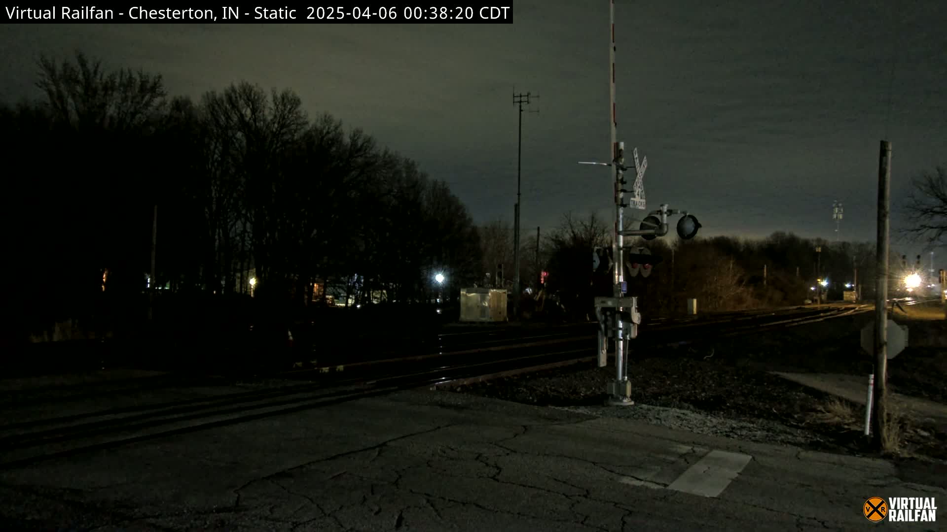 A nighttime view of a railroad crossing with train signals and tracks, surrounded by dark trees under a cloudy sky.