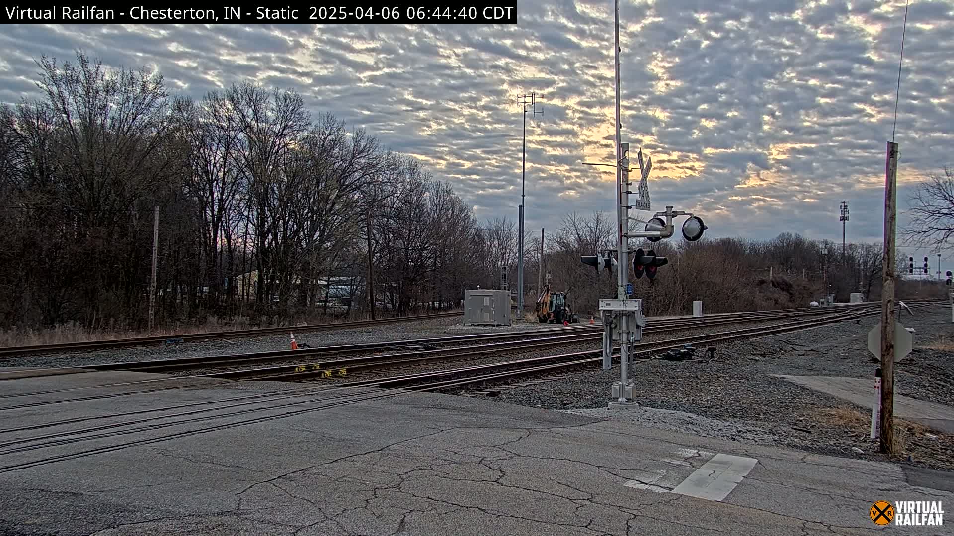 A railroad crossing with multiple tracks is shown under a partly cloudy sky at dawn or dusk, with bare trees and some construction equipment visible in the background.