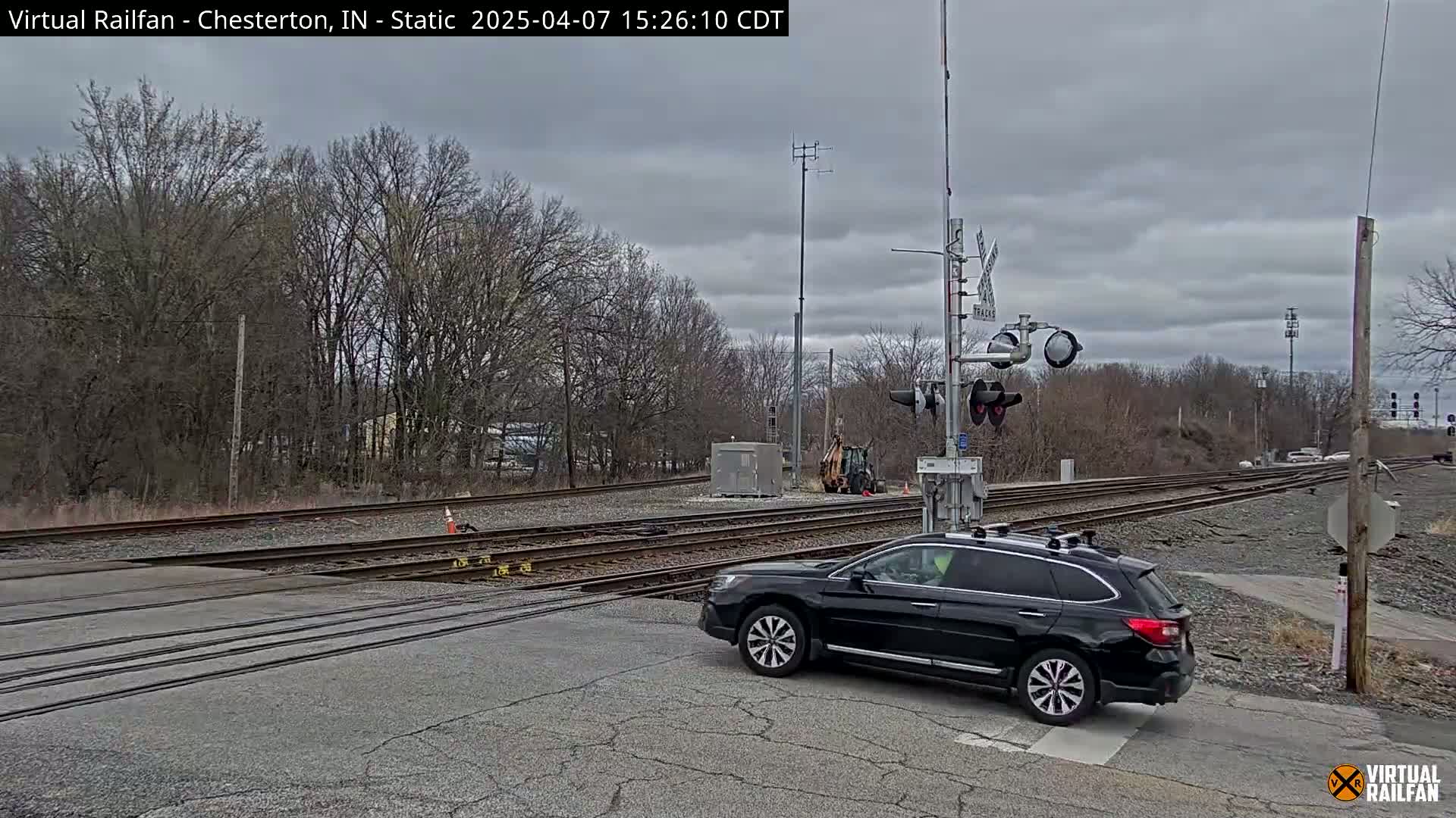 A black SUV waits at a railroad crossing under a cloudy sky, with bare trees and railroad tracks visible in the background.