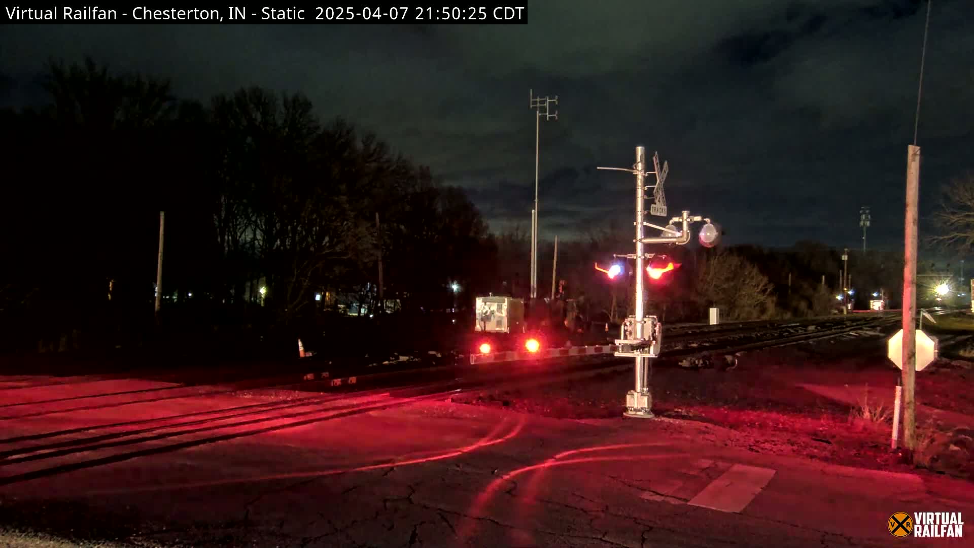 A nighttime view of a railroad crossing with flashing red lights and crossing gates down, illuminated by the red lights against a dark background of trees under a cloudy sky.