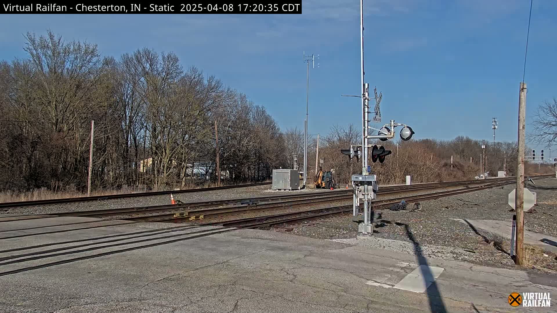 A sunny day reveals a railroad crossing with several tracks, railway signals, a small piece of construction equipment, and bare trees in the background.