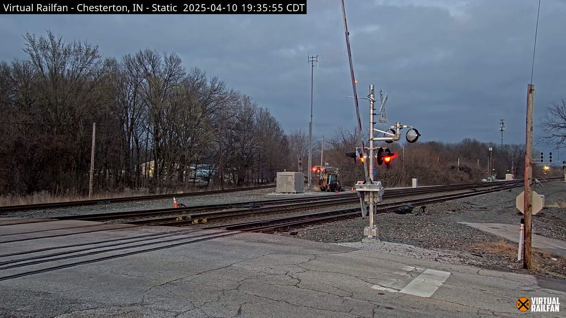 A railroad crossing with lowered gates and flashing red lights is shown under a cloudy, twilight sky, with bare trees and a small piece of construction equipment visible in the background.