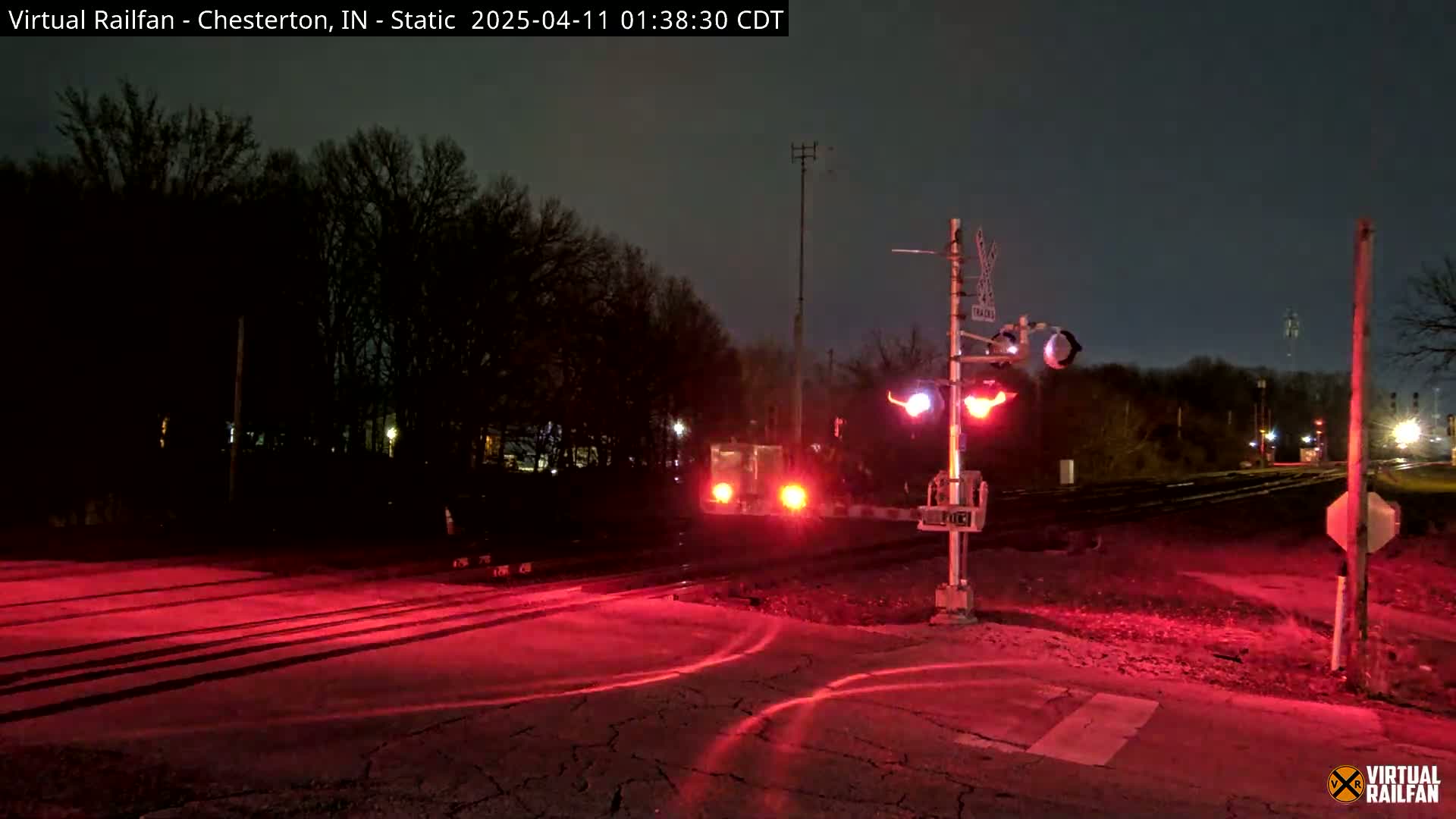 At night, a train approaches a railroad crossing illuminated by red lights, casting a red glow on the surrounding area.