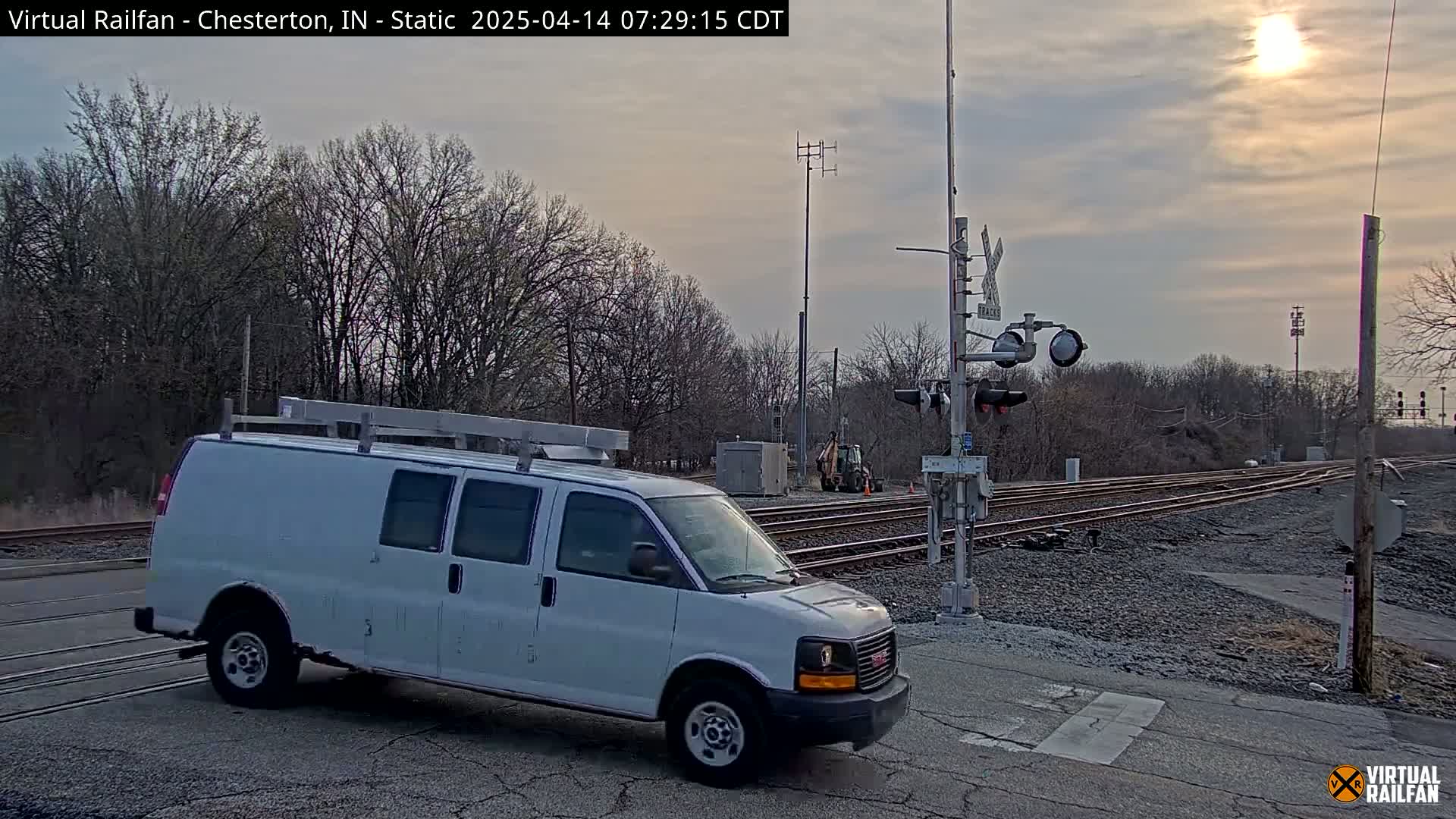 A white cargo van is stopped at a railroad crossing under a partly cloudy sky, with the sun visible.