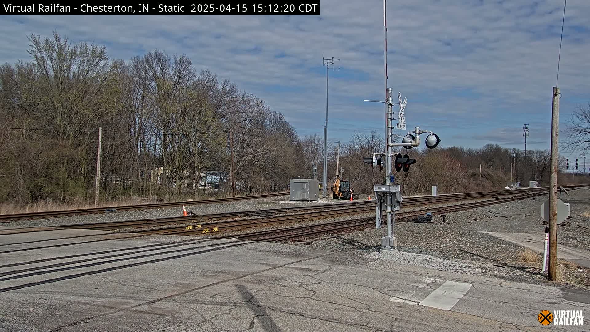 A railroad crossing with a partly cloudy sky shows several train tracks intersecting a road, with bare trees in the background and a small piece of construction equipment visible.