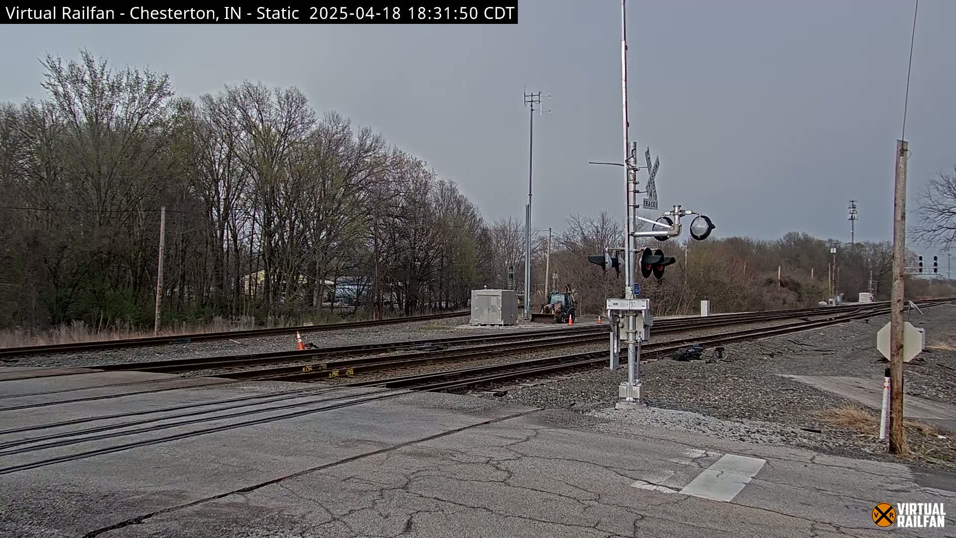 A railroad crossing with multiple tracks is shown under an overcast sky, surrounded by bare trees.