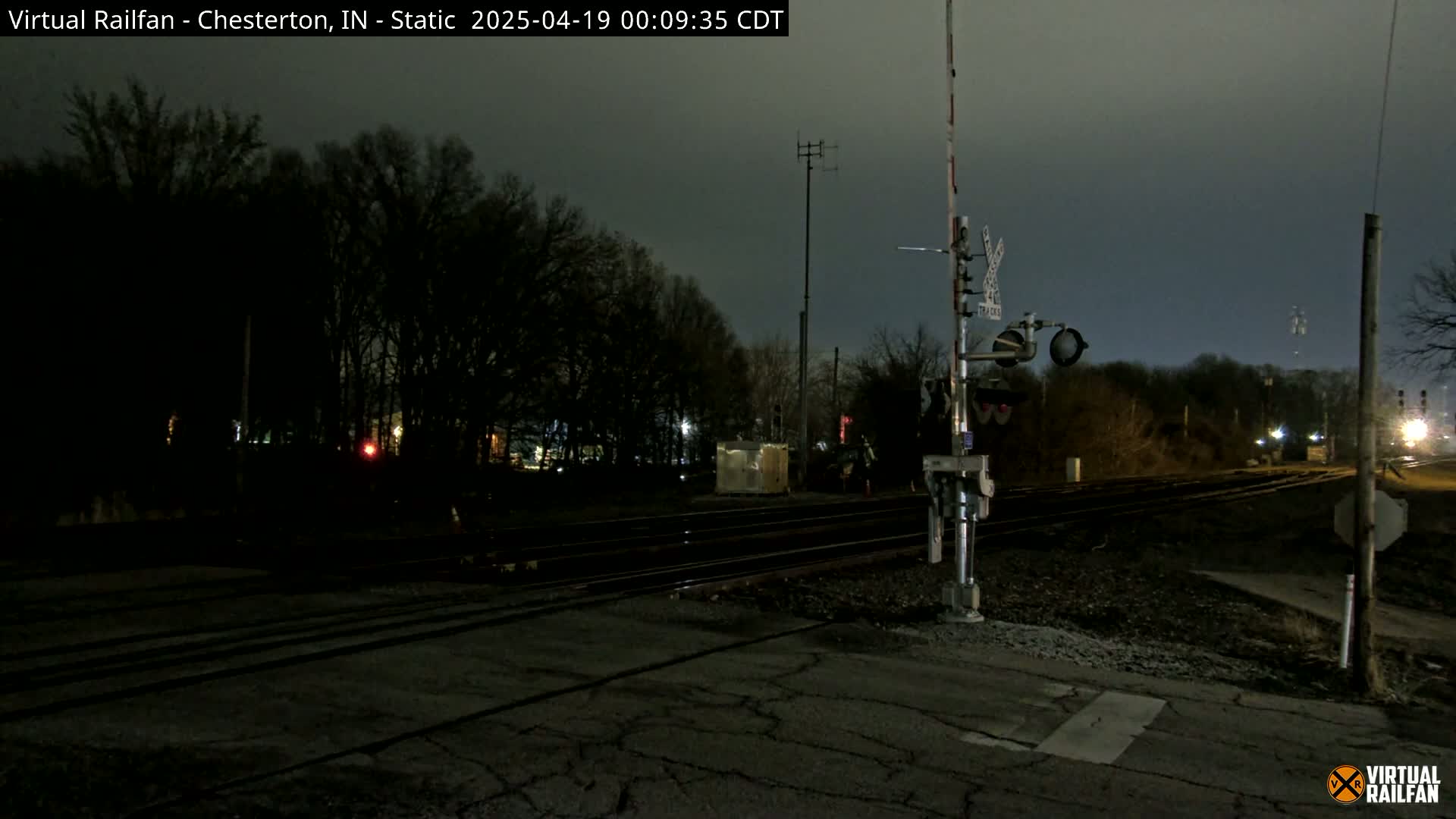 A nighttime view of a railroad crossing with train tracks, crossing signals, and bare trees under a dark, overcast sky.