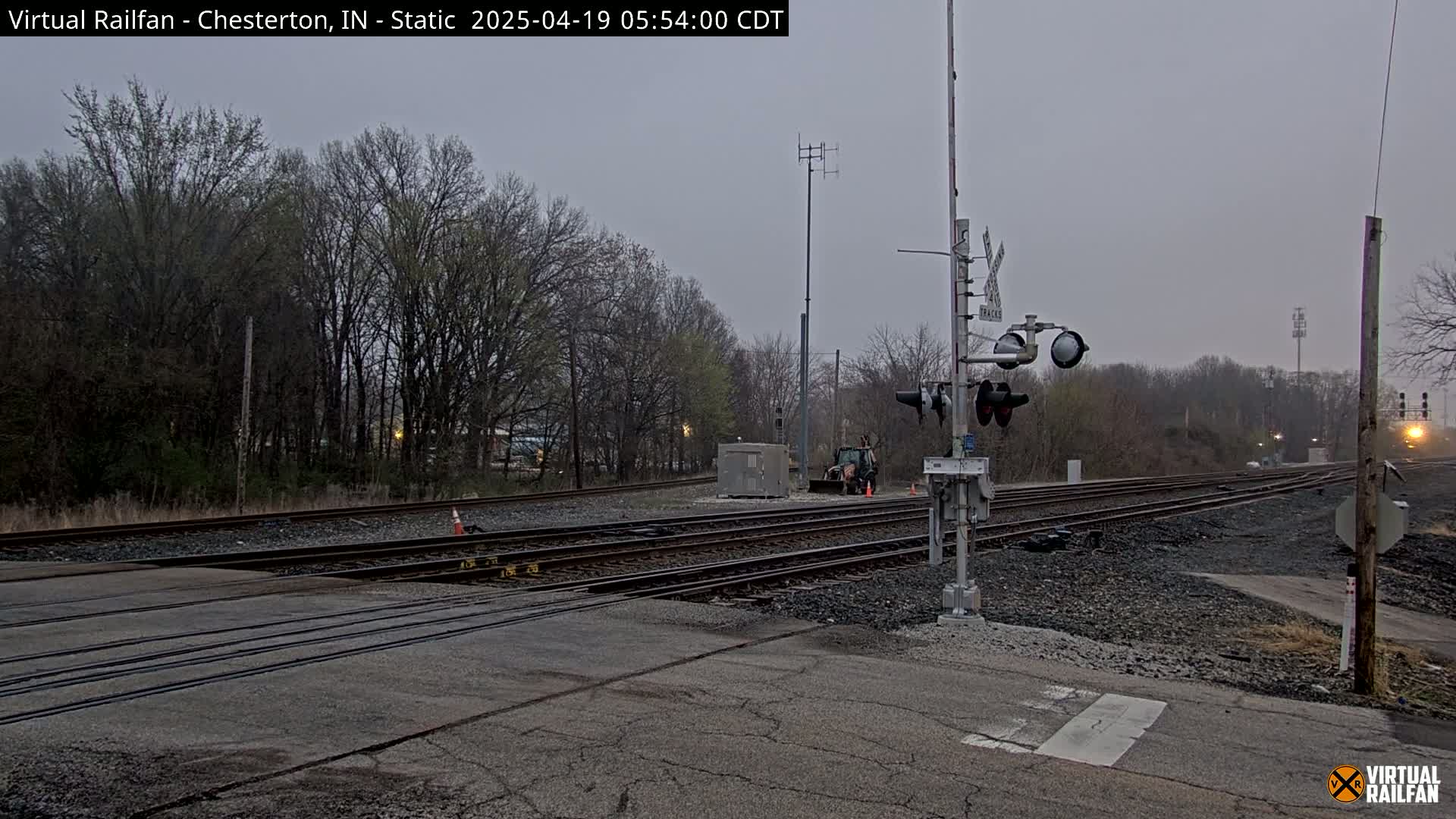 A railroad crossing with a train signal and multiple tracks is visible under an overcast sky, with bare trees lining the background.