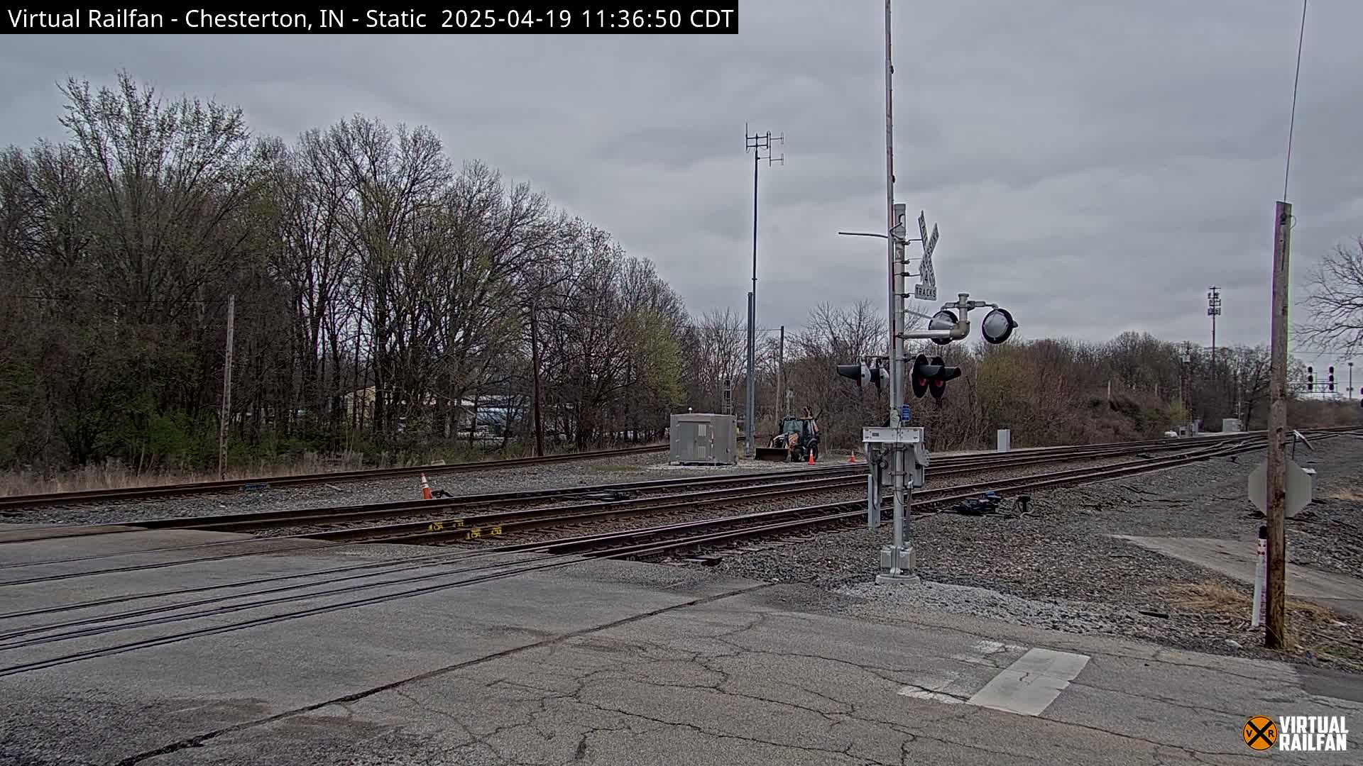 A railroad crossing with multiple tracks is shown under an overcast sky, surrounded by bare trees and some early spring vegetation.
