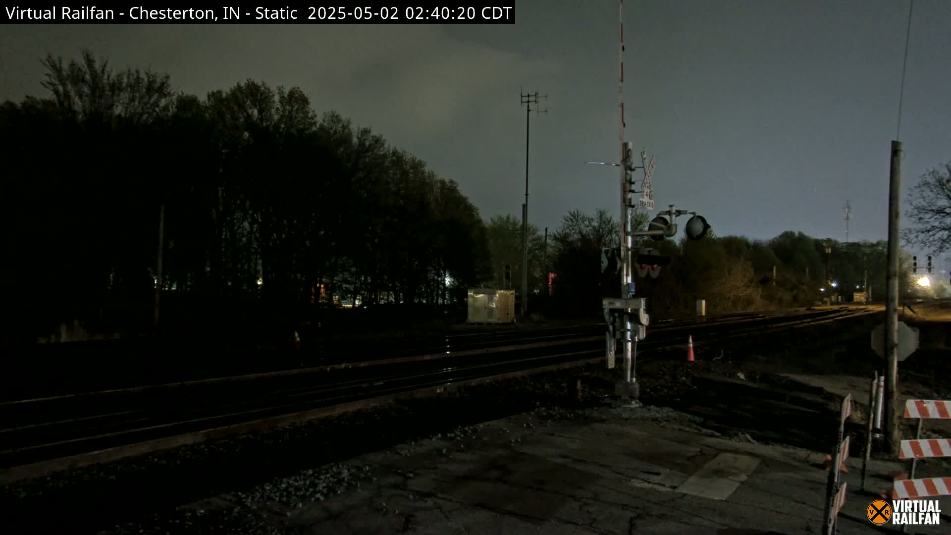 A nighttime view of a railroad crossing with train tracks extending into a dark, tree-lined area under a cloudy sky.