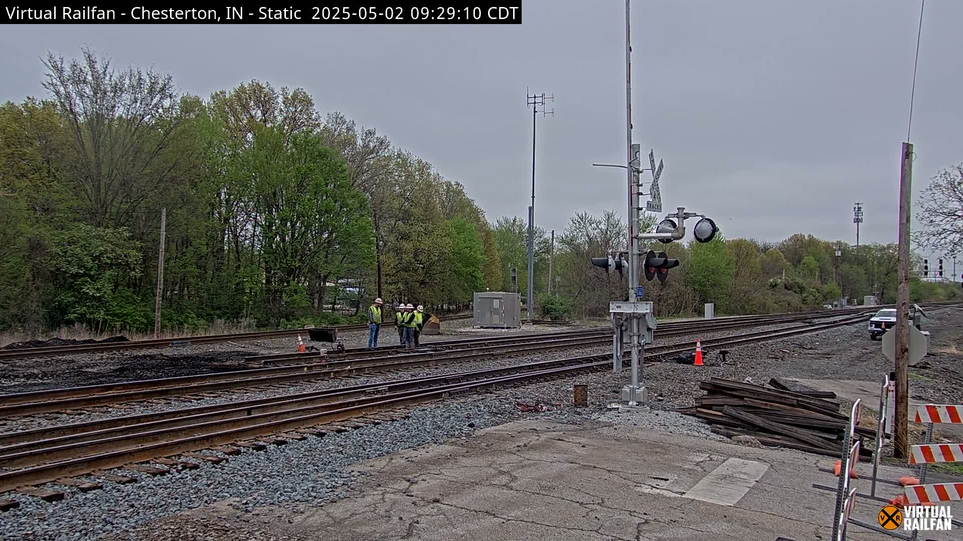Under an overcast sky, several workers stand near railroad tracks undergoing some form of maintenance or construction, with trees lining the background.