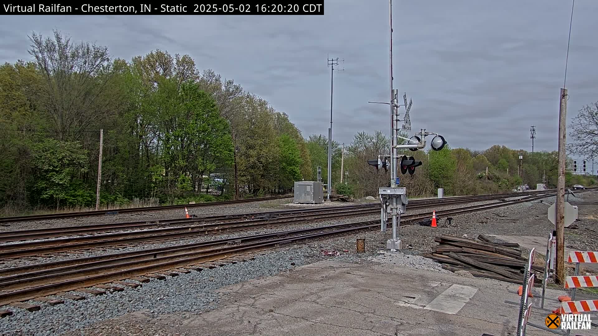 Multiple railroad tracks extend into a wooded area under an overcast sky, with railroad crossing signals and construction materials visible near the foreground.