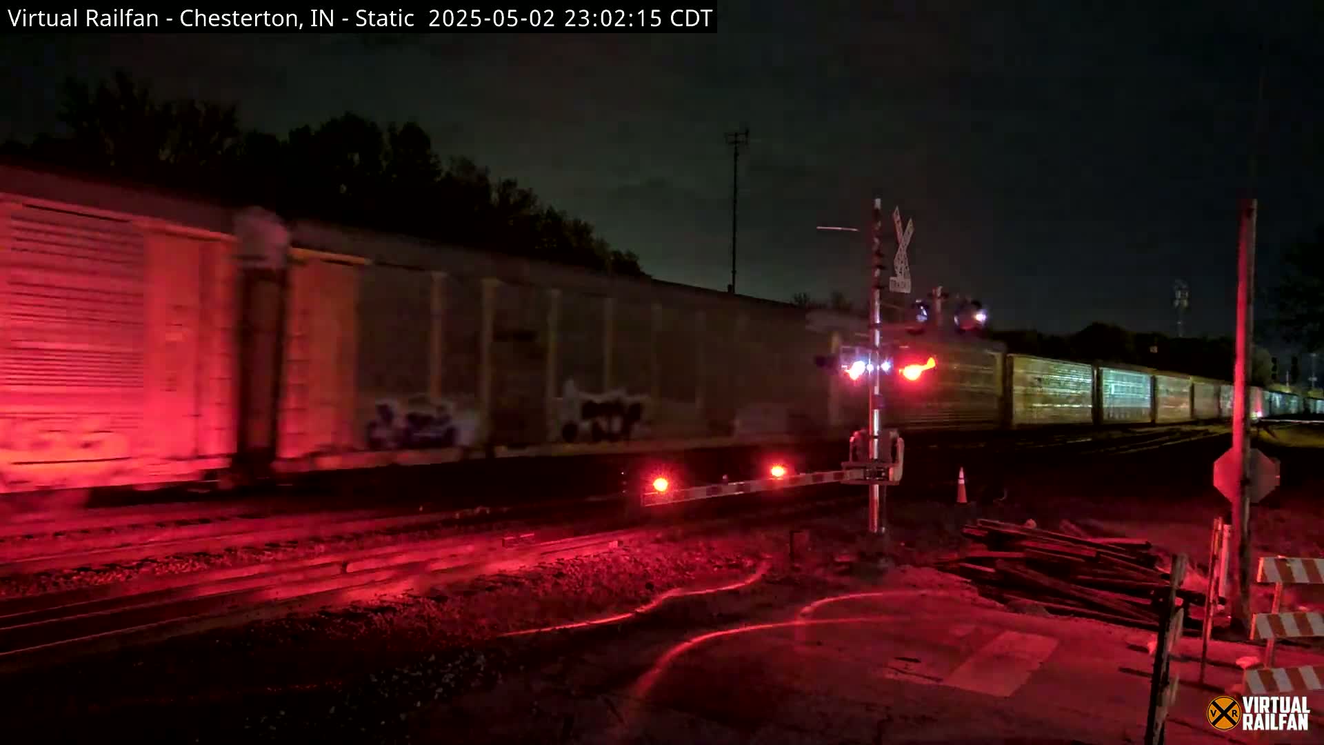 A long freight train passes a railway crossing at night, illuminated by red lights from the crossing gates and the train itself.