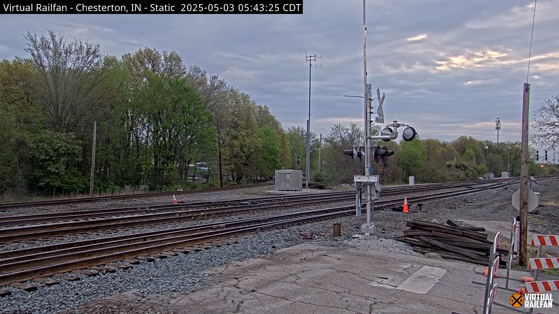 A railroad crossing with multiple tracks under an overcast sky, surrounded by trees and some construction materials.