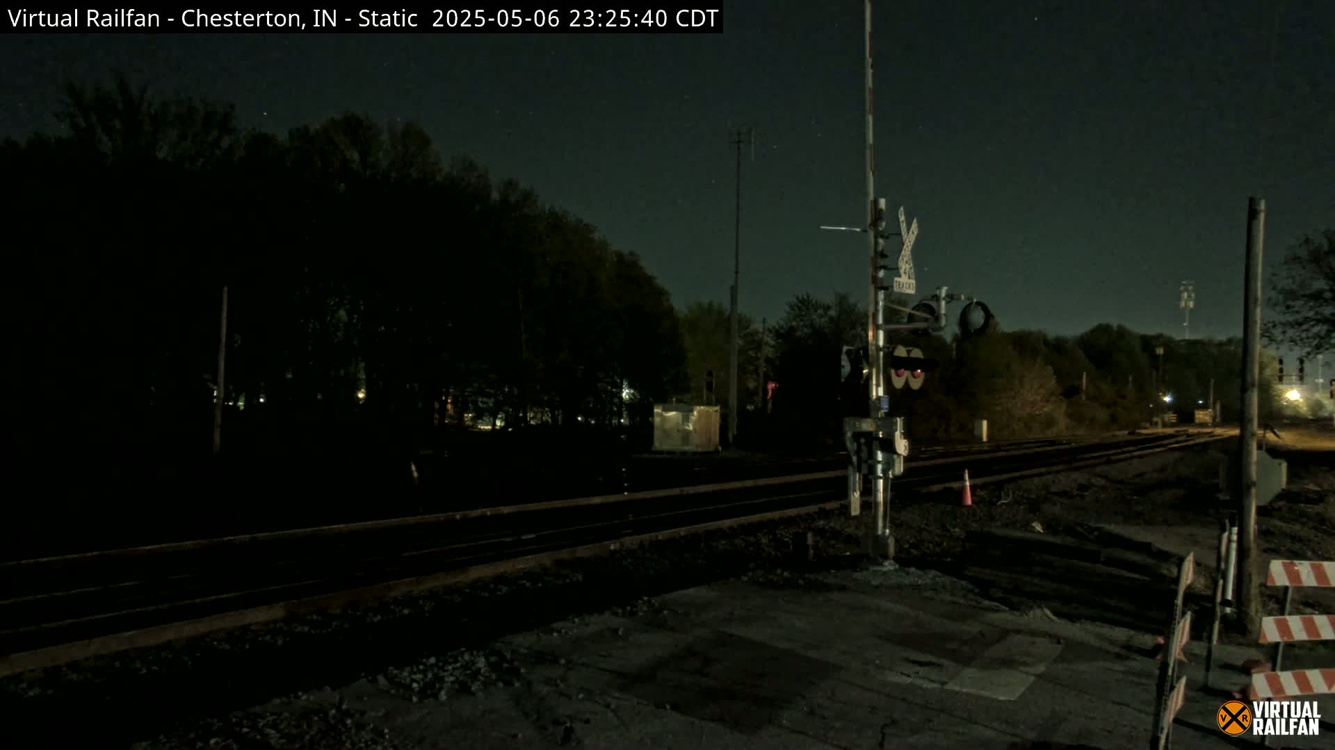 Under a dark, nighttime sky, a set of railroad tracks stretches into the distance, bordered by trees, with a railway crossing signal visible in the foreground.