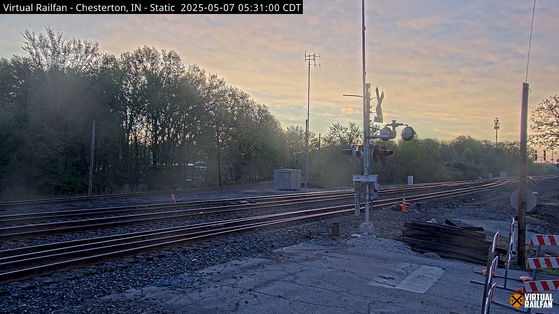 Multiple sets of train tracks intersect at a rail yard during a hazy sunrise, with trees and some construction materials visible.