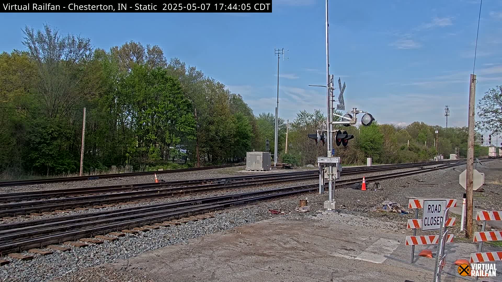 A railroad crossing is closed under a mostly clear sky, with multiple train tracks visible and lush green trees in the background.